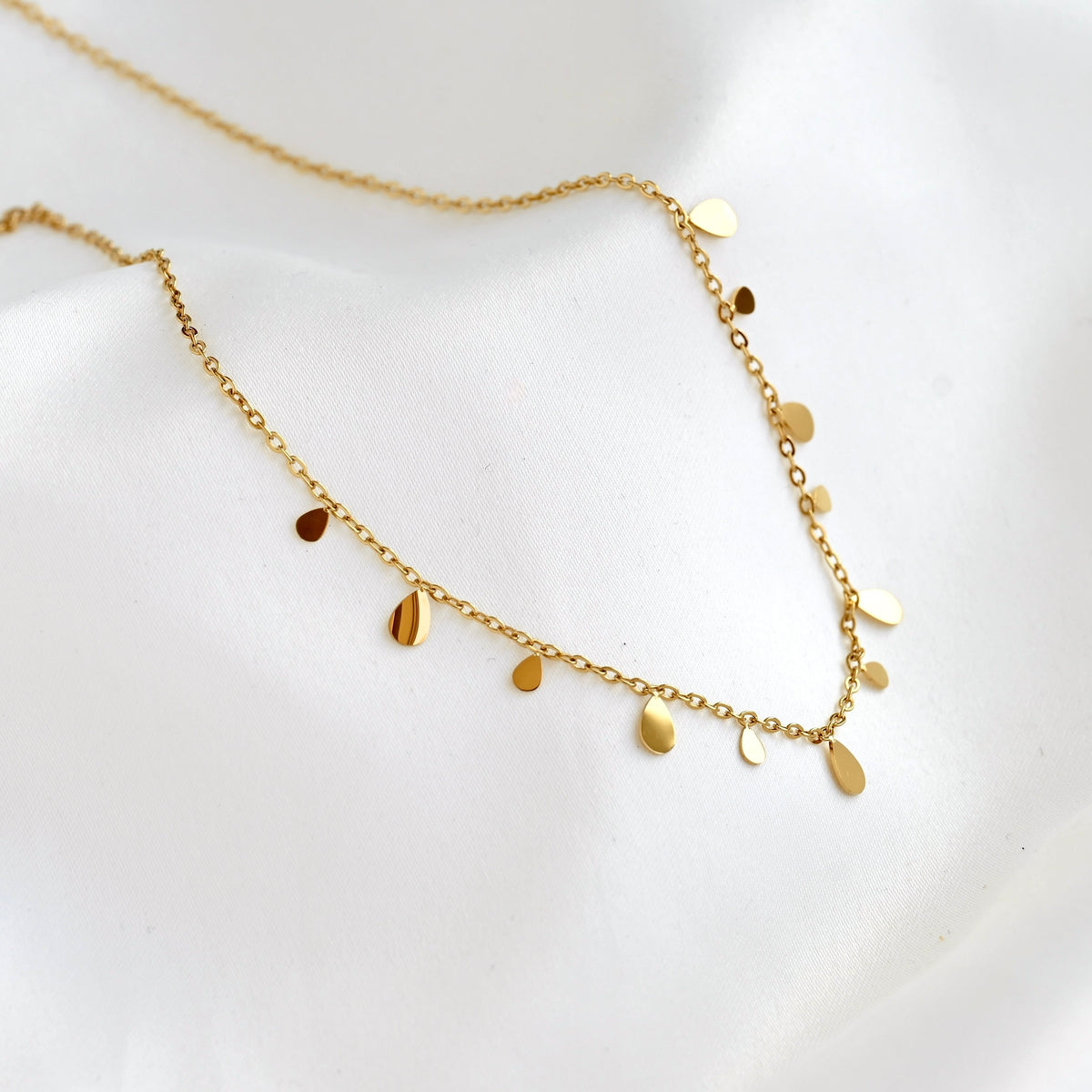 A delicate gold chain necklace with small flat teardrop-shaped discs, displayed on a soft white fabric background.
