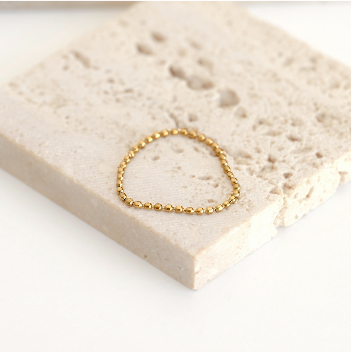 A delicate gold beaded bracelet resting on a textured beige stone tile against a clean white background.