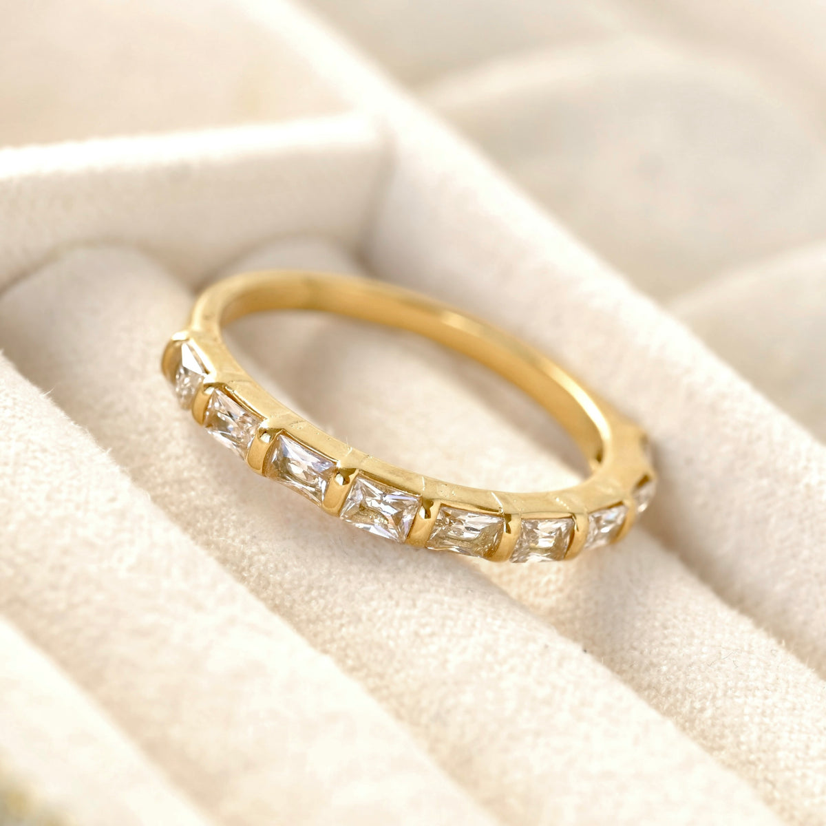 A yellow gold half eternity ring featuring baguette-cut clear stones, resting on a cream-colored velvet ring box.