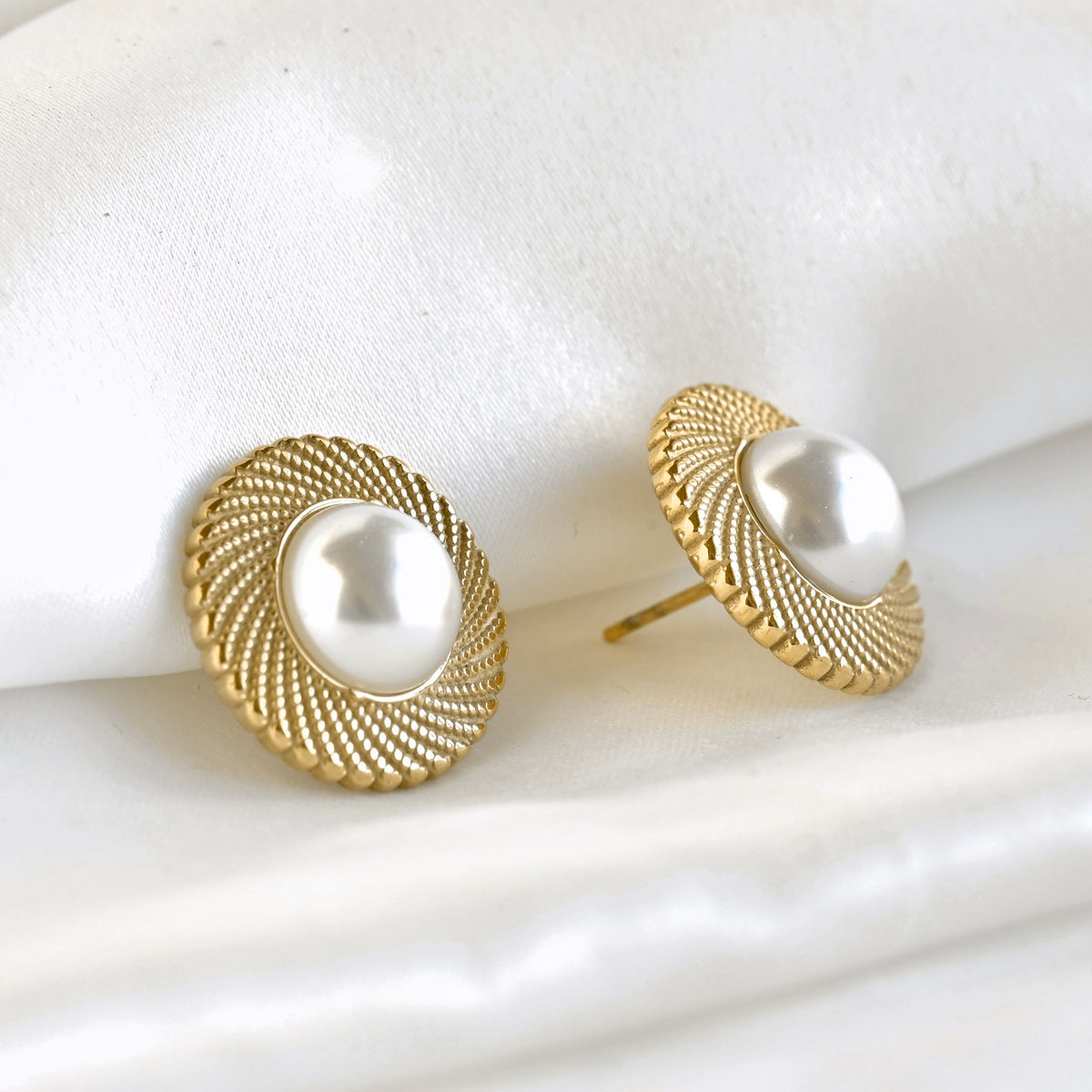 A pair of gold round stud earrings with a textured spiral milgrain border and a central white simulated pearl, displayed on a soft white fabric background.