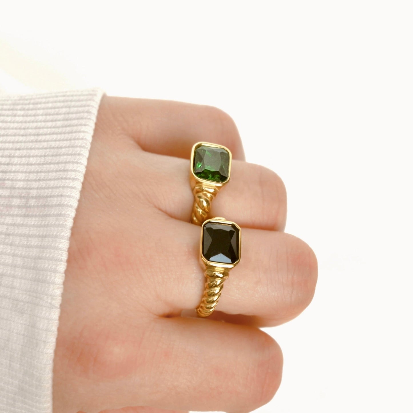 Two gold rings with square green gemstones worn on a person's fingers, shown against a clean white background with a white ribbed sleeve visible on the left.