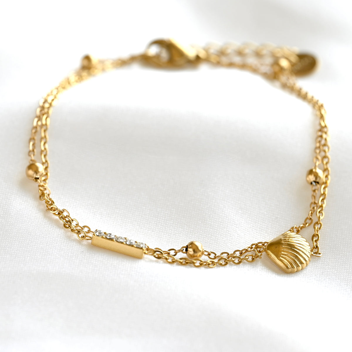 A delicate gold chain bracelet with a lobster clasp, featuring a small seashell charm, a bar with tiny clear stones, and small gold beads, displayed on a white fabric background.