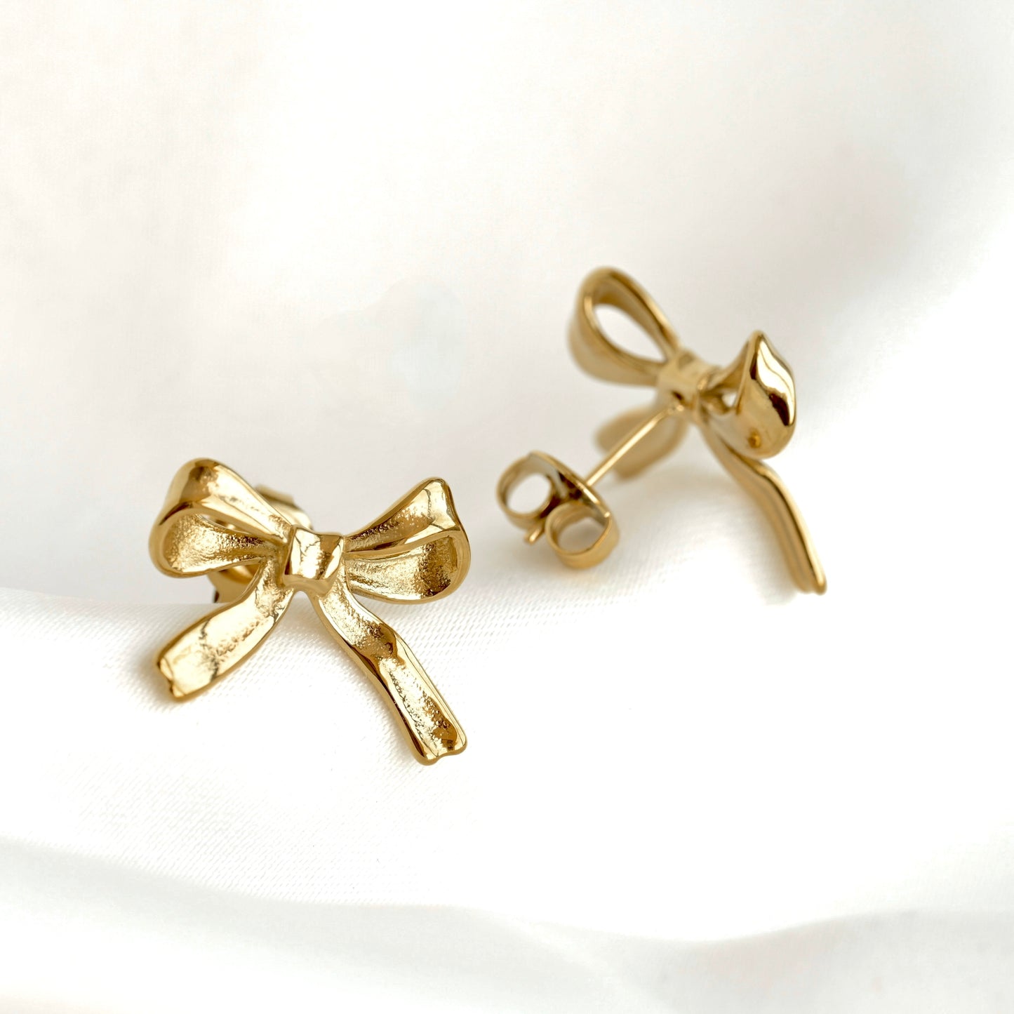 A pair of gold bow stud earrings, with one shown from the front and the other from the side/back to display the post and backing, resting on a white fabric background.