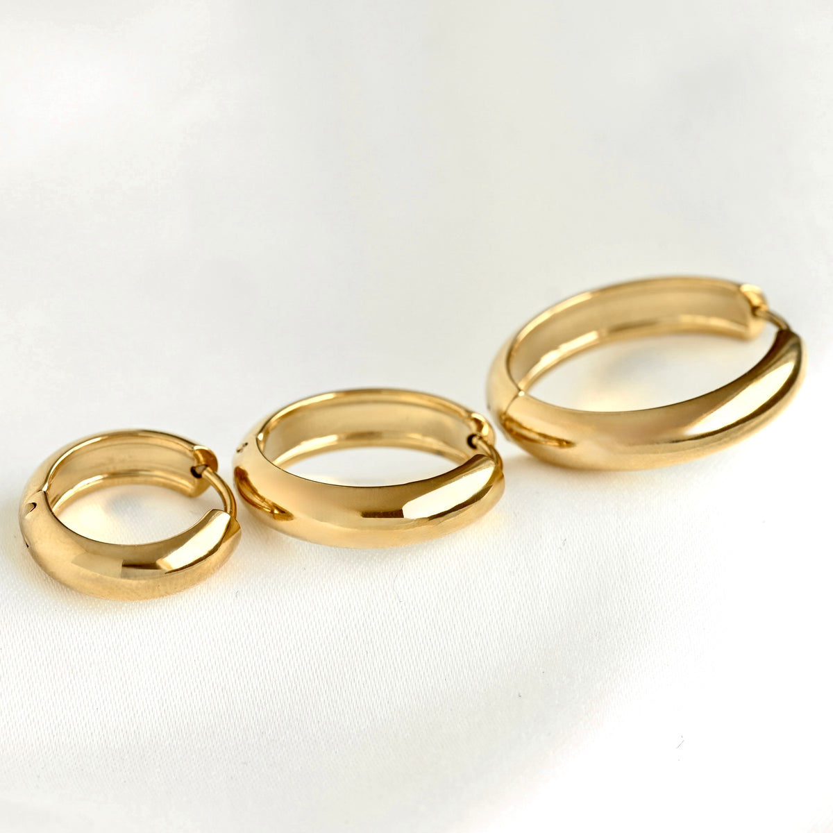 Three gold-plated thick hoop earrings in different sizes, arranged diagonally on a clean white textured fabric background.