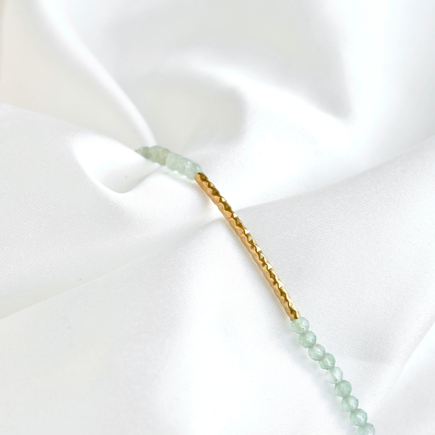 A delicate gold bracelet with light green faceted bead accents and round hammered bar in 18k gold PVD stainless steel displayed on a draped white satin fabric.