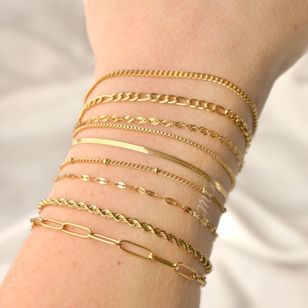 Gold paperclip, rope, mariner, satellite, herringbone, box, singapore, figaro and curb bracelets on a wrist against a neutral background