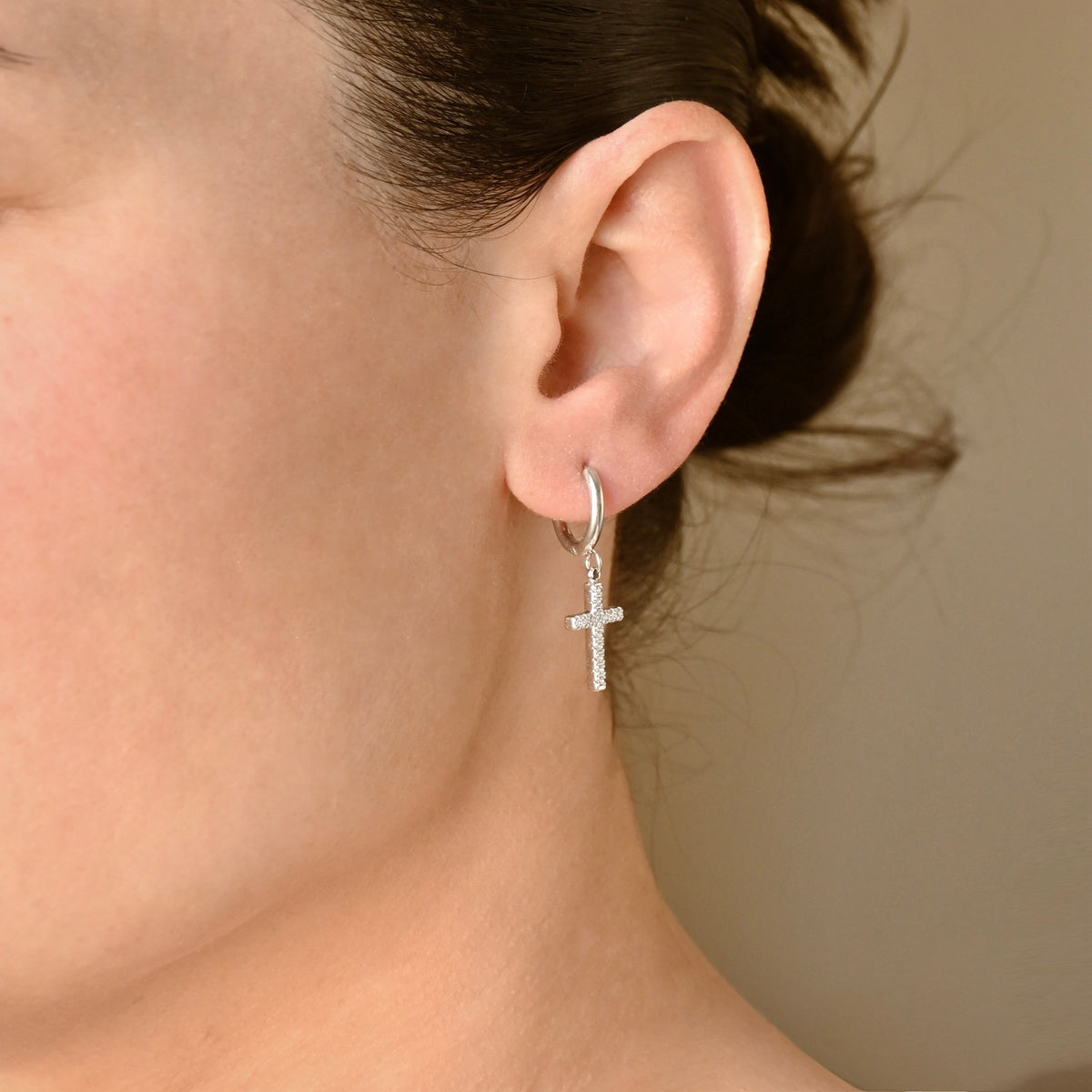 A close-up of a person's ear wearing a small silver hoop earring with a diamond cross pendant, shown against a plain beige background.