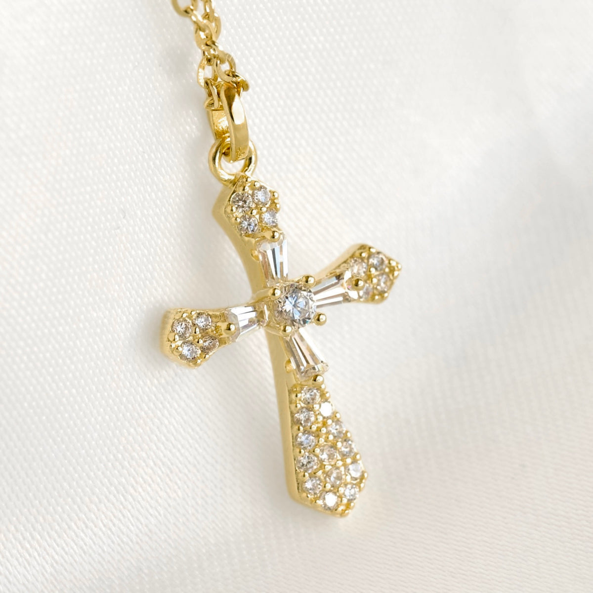 A gold cross pendant necklace adorned with clear crystals, shown on a white textured fabric background.