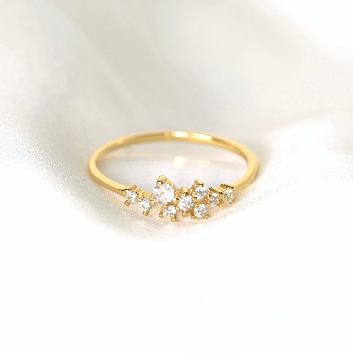 A delicate gold ring featuring a small cluster of clear round-cut CZ stones in a floral or leaf-like arrangement, resting on a soft white fabric background.