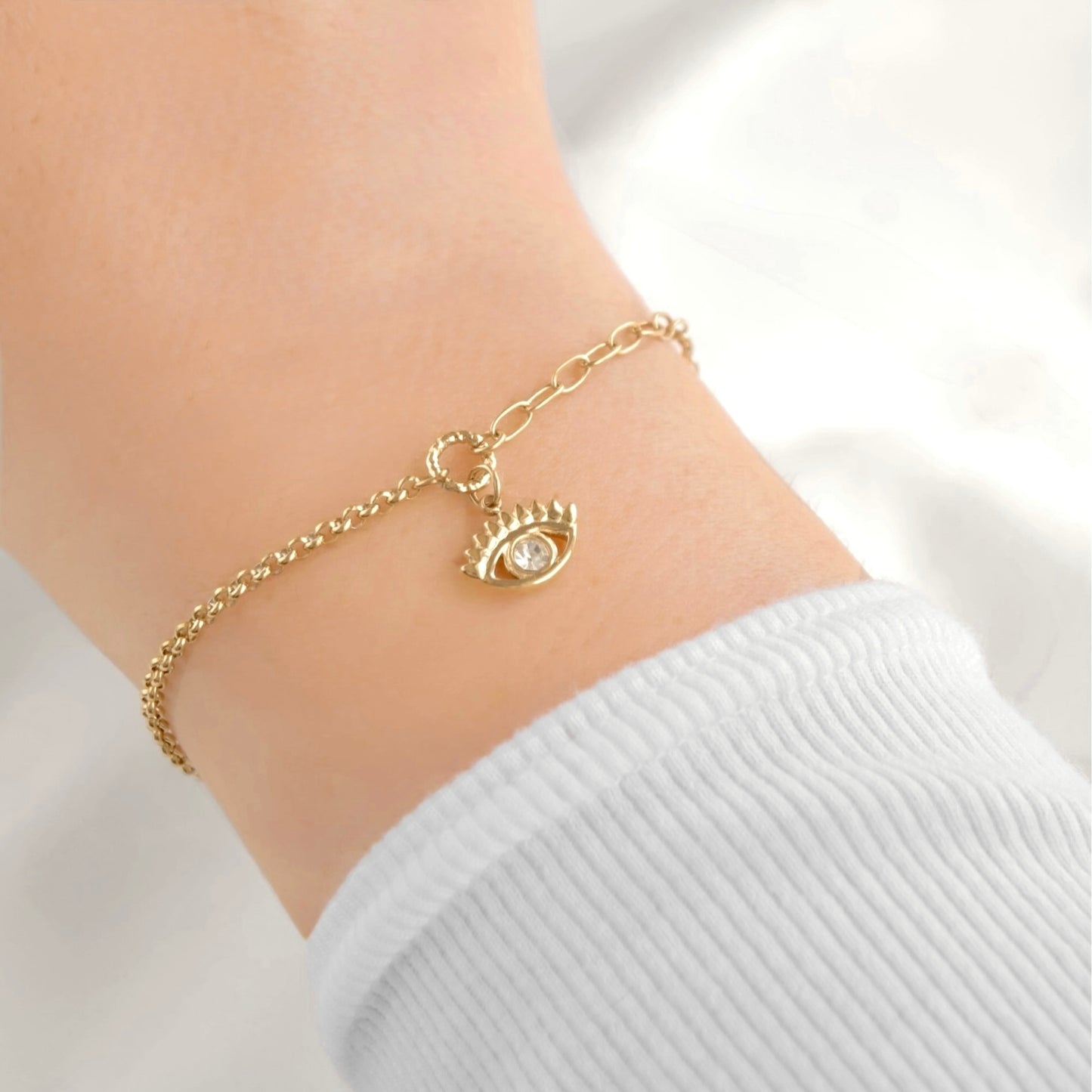 A delicate gold chain bracelet with an evil eye charm, shown worn on a person's wrist over a white ribbed long-sleeve top.