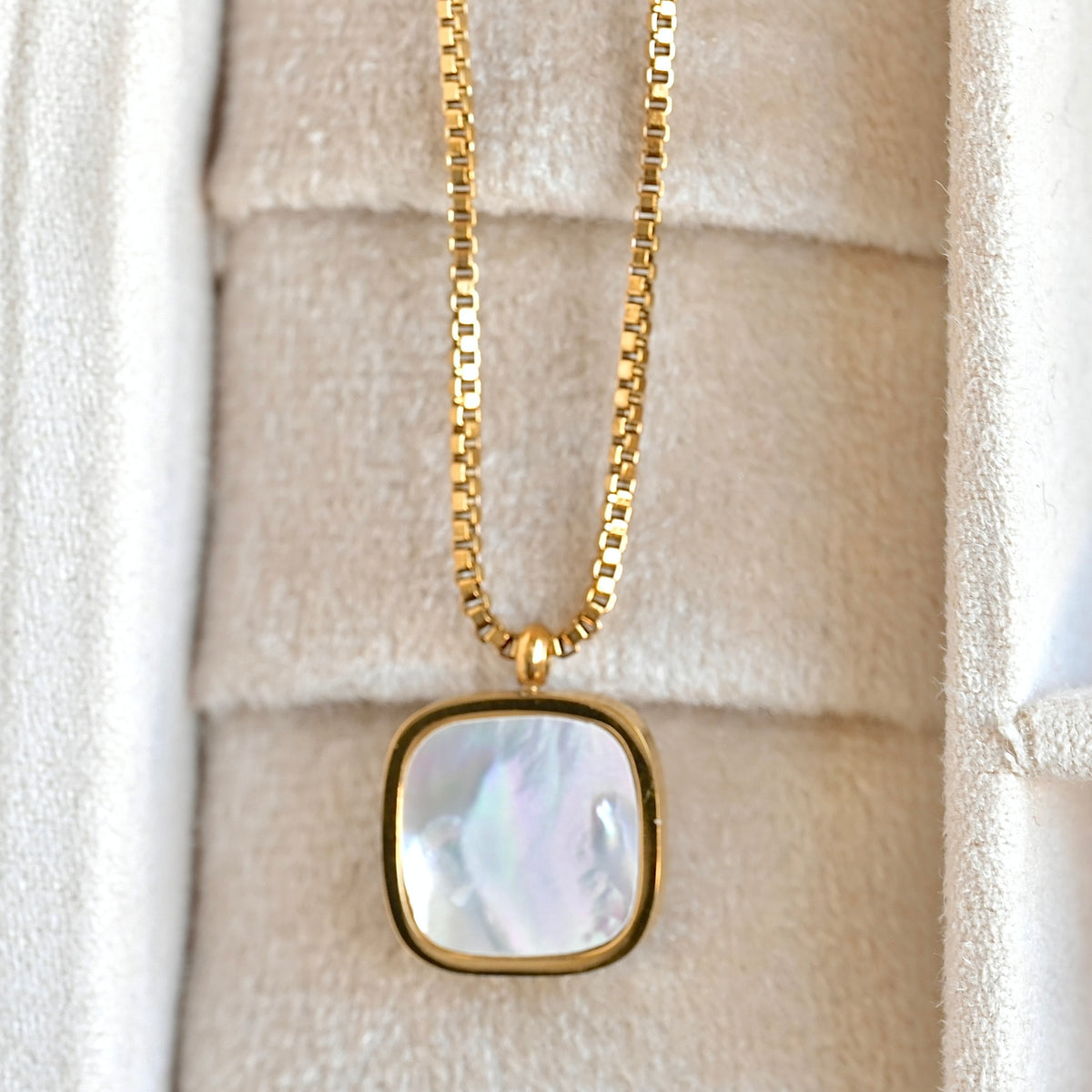 A gold box chain necklace with a square pendant featuring a pearlescent white and iridescent shell inlay, displayed inside a cream-colored jewelry box.