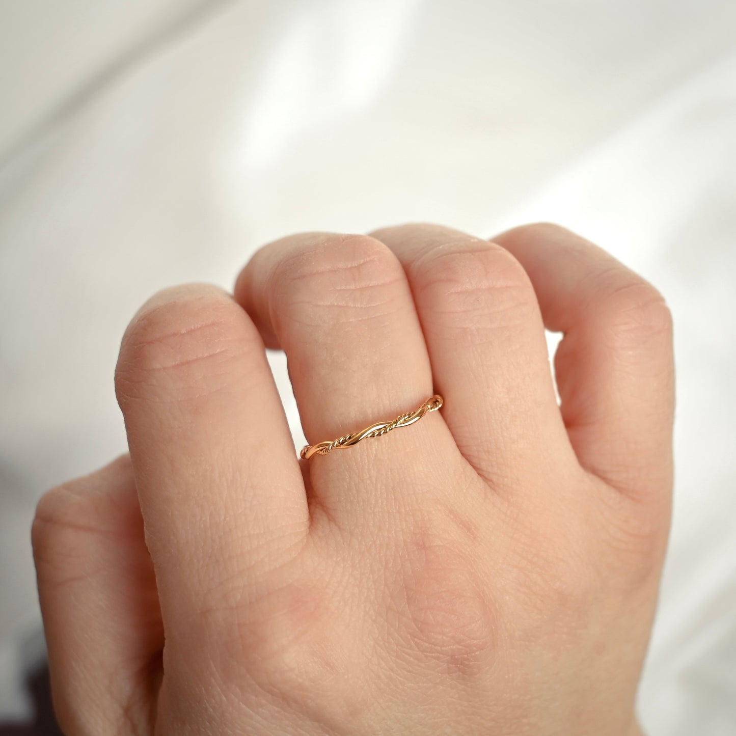 A delicate gold ring with a twisted, rope-like design worn on the ring finger of a hand, shown in a close-up shot against a soft, light-colored background.