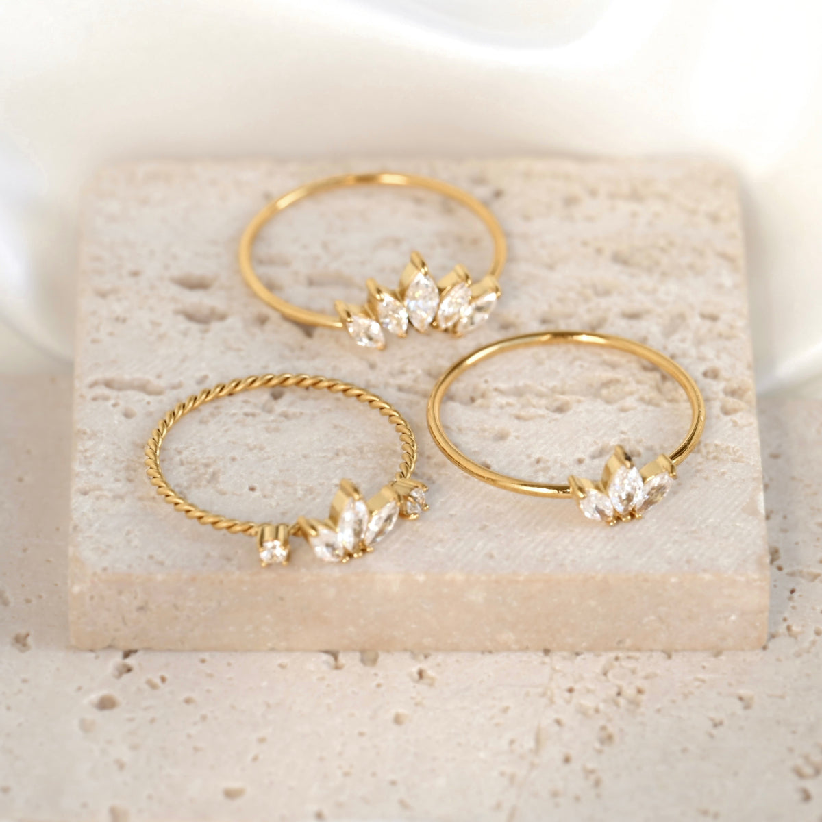 Three gold rings displayed on a light stone slab. Two rings are smooth bands with small clear crystal leaf clusters, and one ring is twisted with a matching crystal leaf cluster.