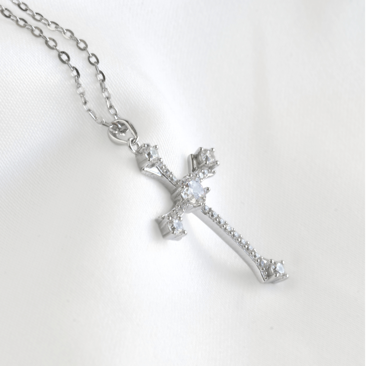 A silver cross pendant necklace adorned with clear crystals, resting on a white satin fabric background.