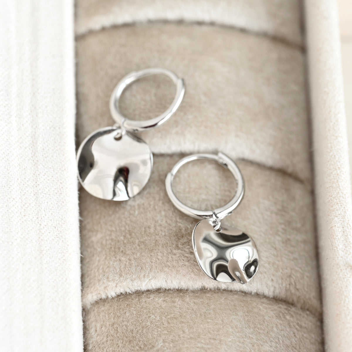 A pair of polished silver hoop earrings with dangling hammered circle charms, displayed on a beige velvet jewelry box.