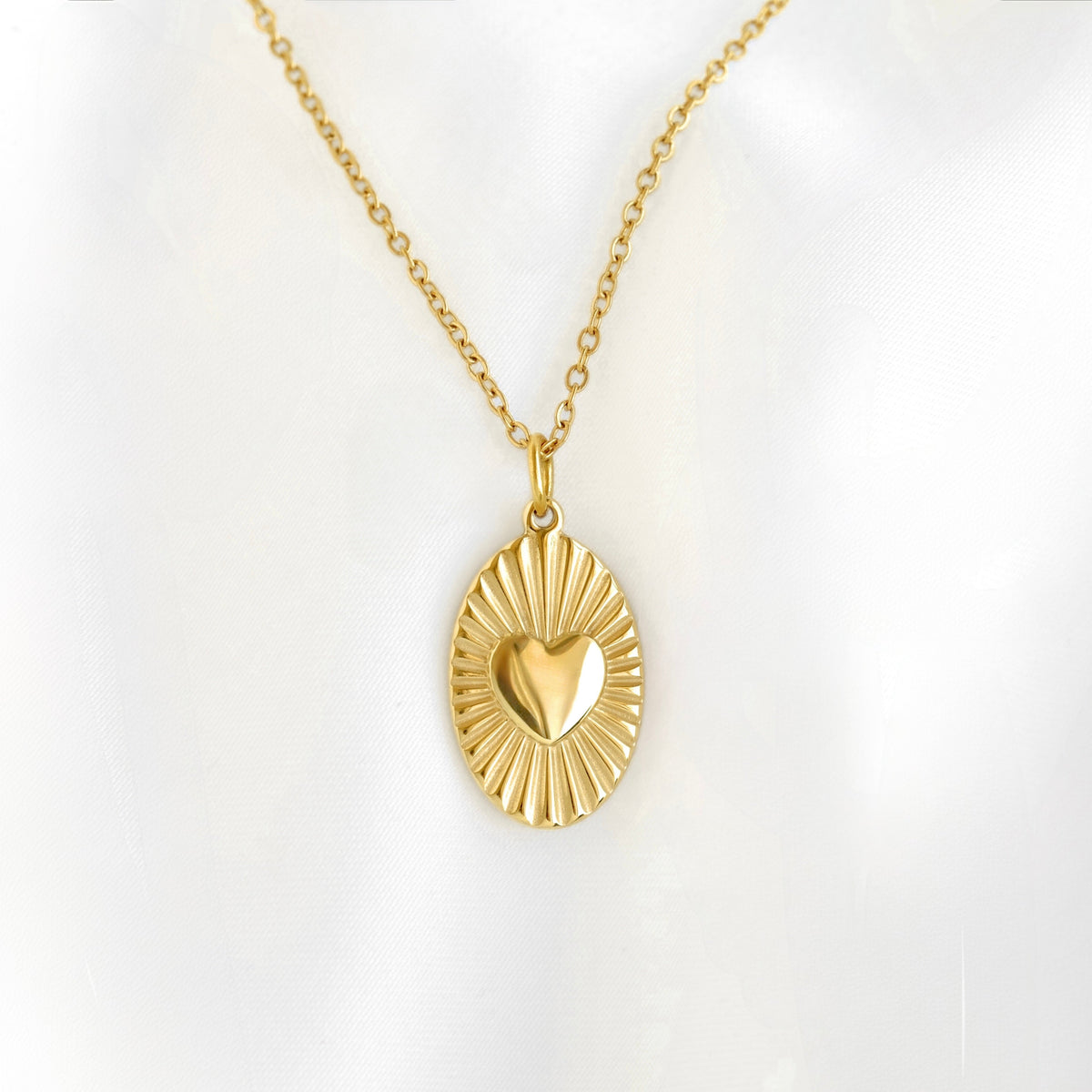 A gold pendant necklace featuring a sunburst oval design with a polished heart at the center, displayed on a soft white background.