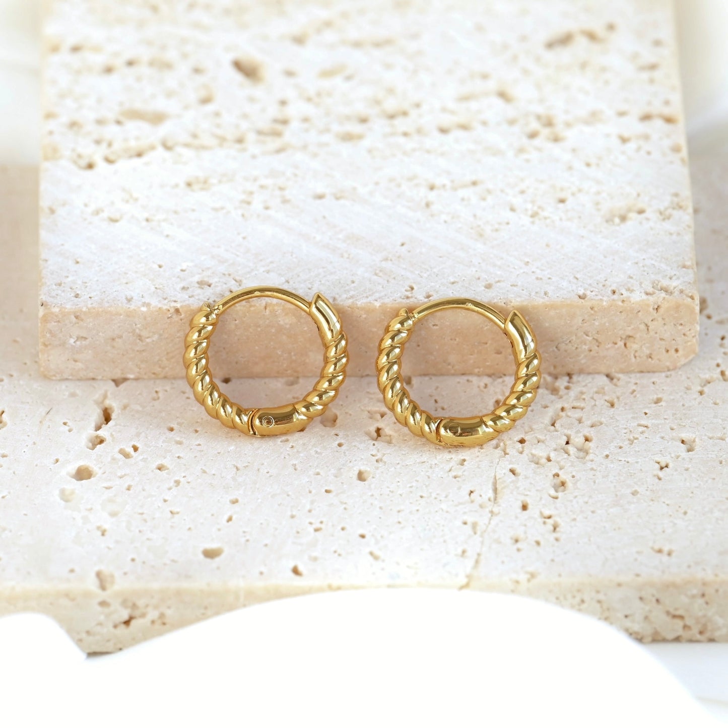 A pair of small twisted hoop earrings, displayed on a light beige stone tile.