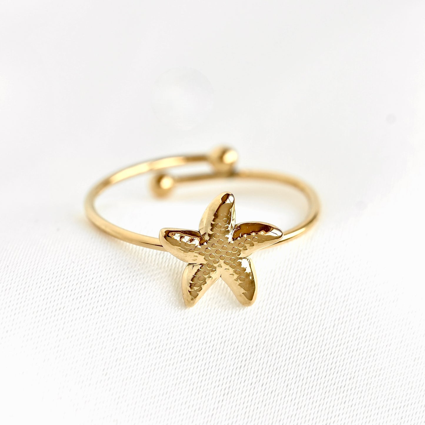 Front view of a gold plated adjustable sea star band featuring a textured starfish charm in the center, resting on a white textured fabric background.