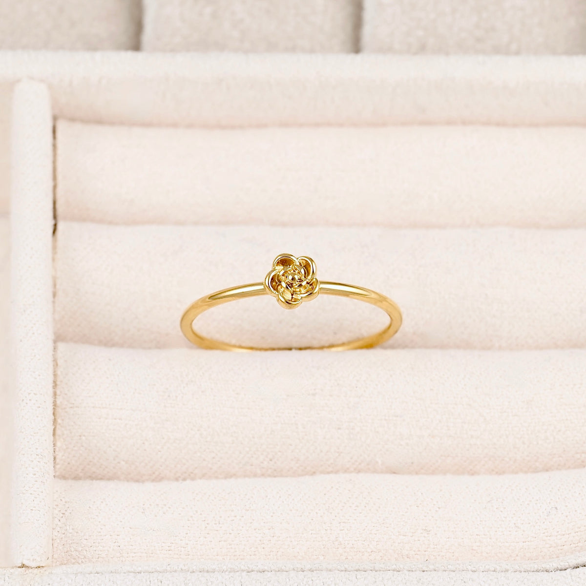 A delicate yellow gold ring featuring a small, intricate floral or knot-like top, displayed on a cream-colored textured ring box.