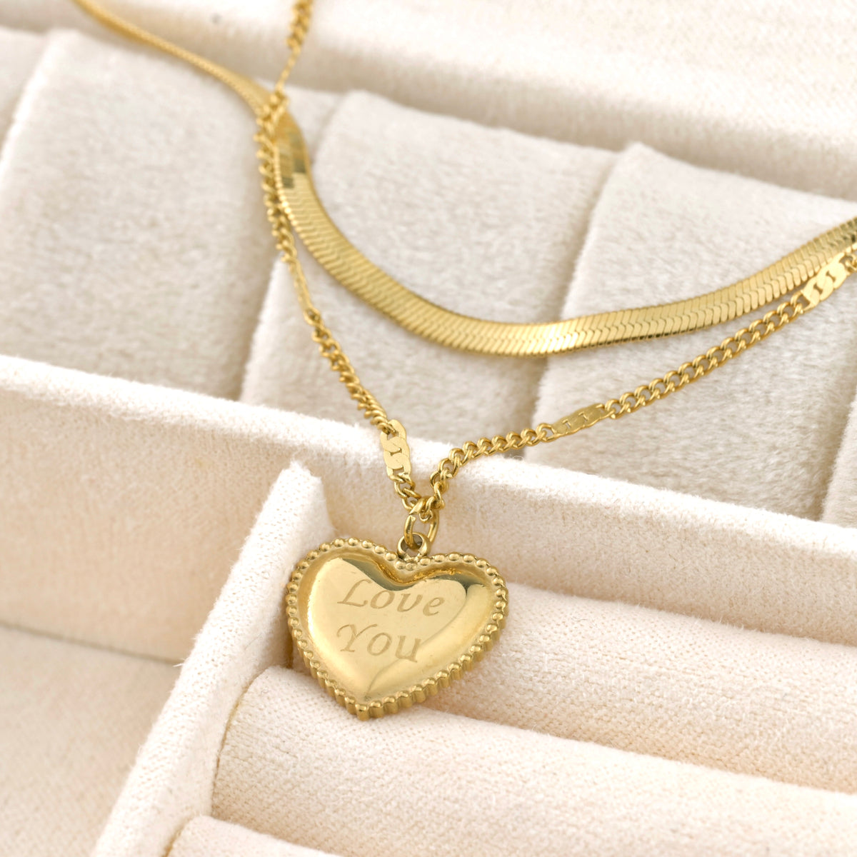 Gold necklace with a heart-shaped pendant that says 'Love You' on a beige cushion.