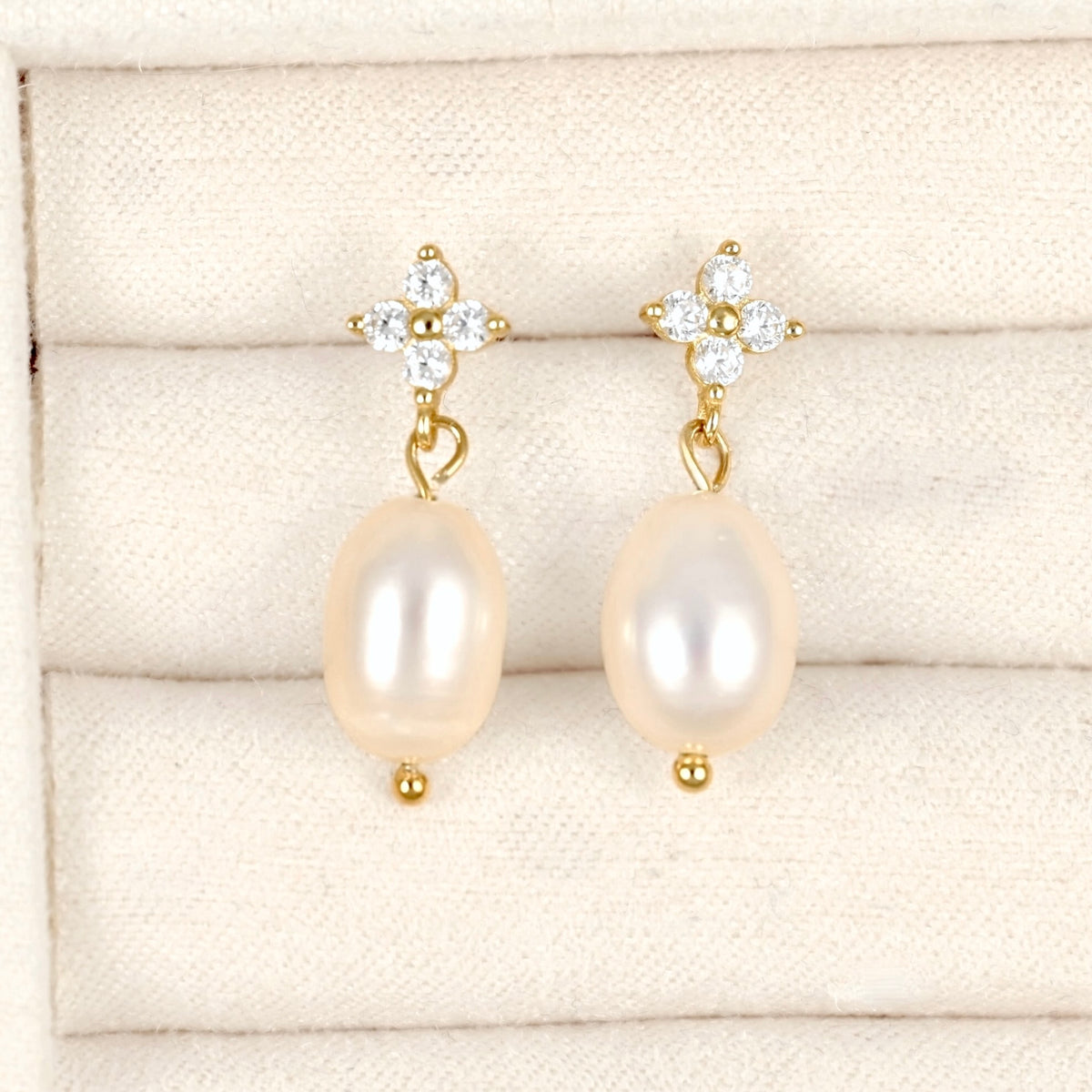 A pair of gold drop earrings with oval cream-colored freshwater pearls and clear crystal floral studs, displayed on a cream-colored textured jewelry box.