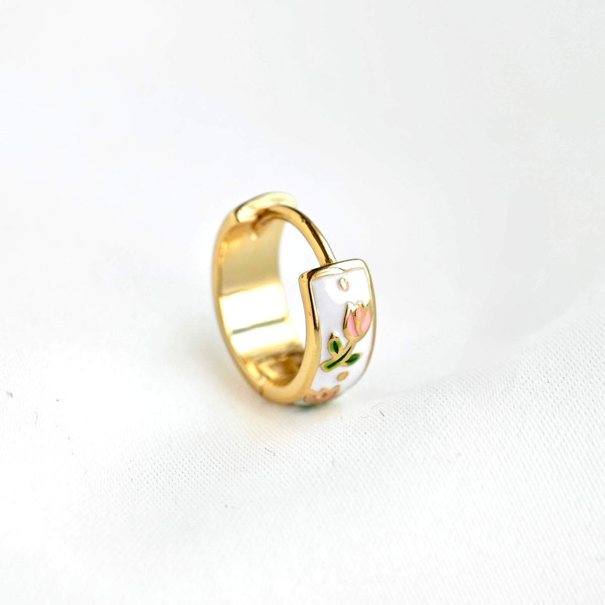 A gold-plated ring with a white enamel inlay featuring a small pink tulip flower and green leaves, displayed on a white textured fabric background.