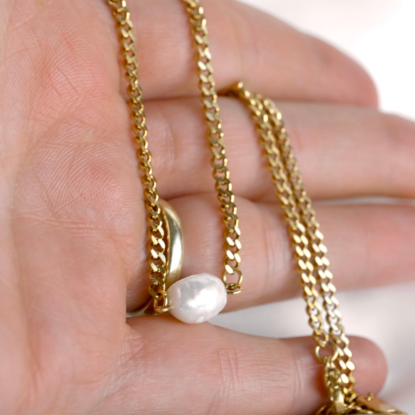 A close-up of a hand holding a gold chain necklace with a small white baroque pearl pendant and a gold signet ring.