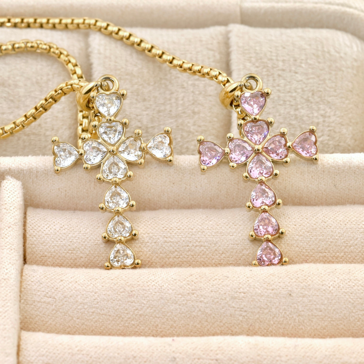 Gold cross pendants with clear and pink heart-cut stones on a beige surface