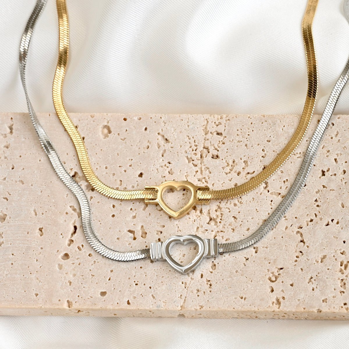 Two snake chain necklaces, one gold and one silver, with a cutout heart charm on a textured surface.
