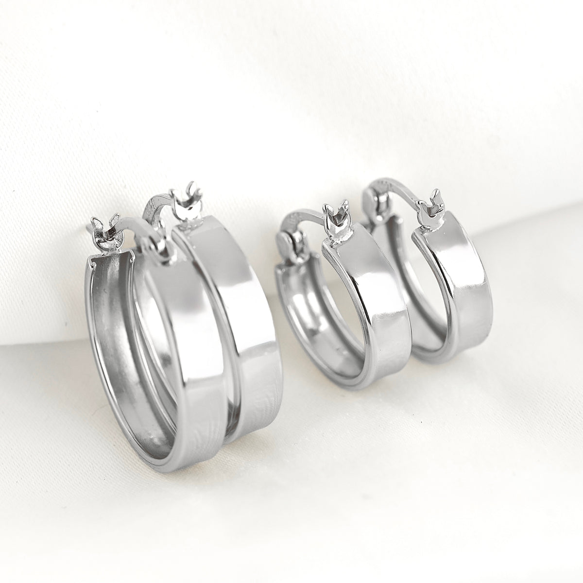 A pair of polished silver-tone hoop earrings with latch back clasp, displayed on a soft white fabric background.