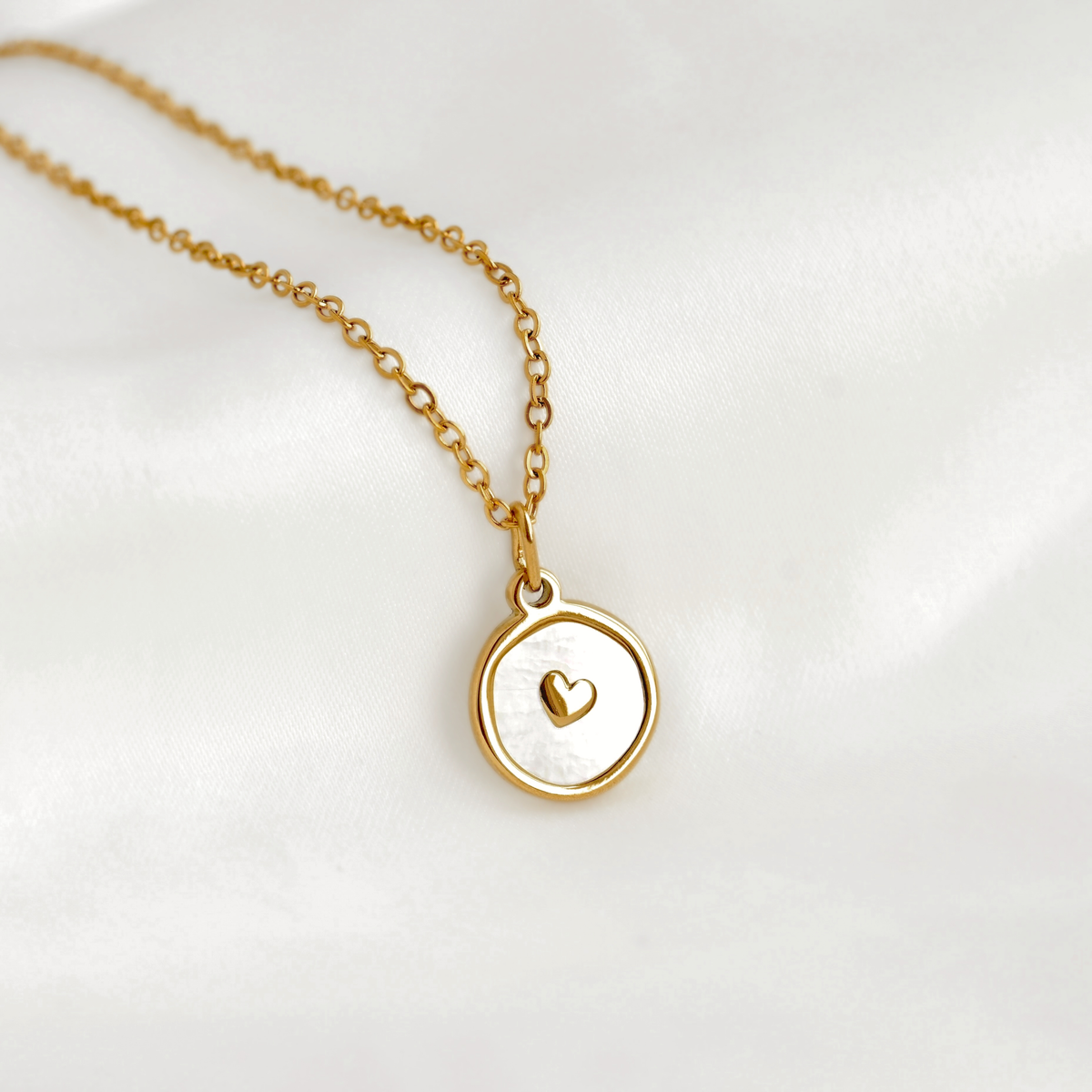A delicate gold chain necklace with a round pendant featuring a white inlay and a small gold heart in the center, displayed on a soft white fabric background.