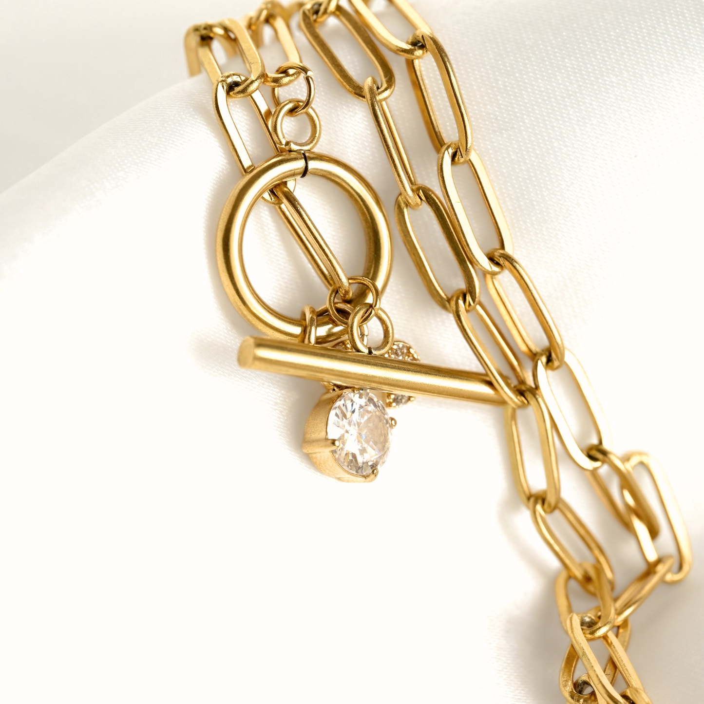 A gold chain bracelet with a toggle clasp and a dangling clear teardrop gemstone, displayed on a white satin surface.