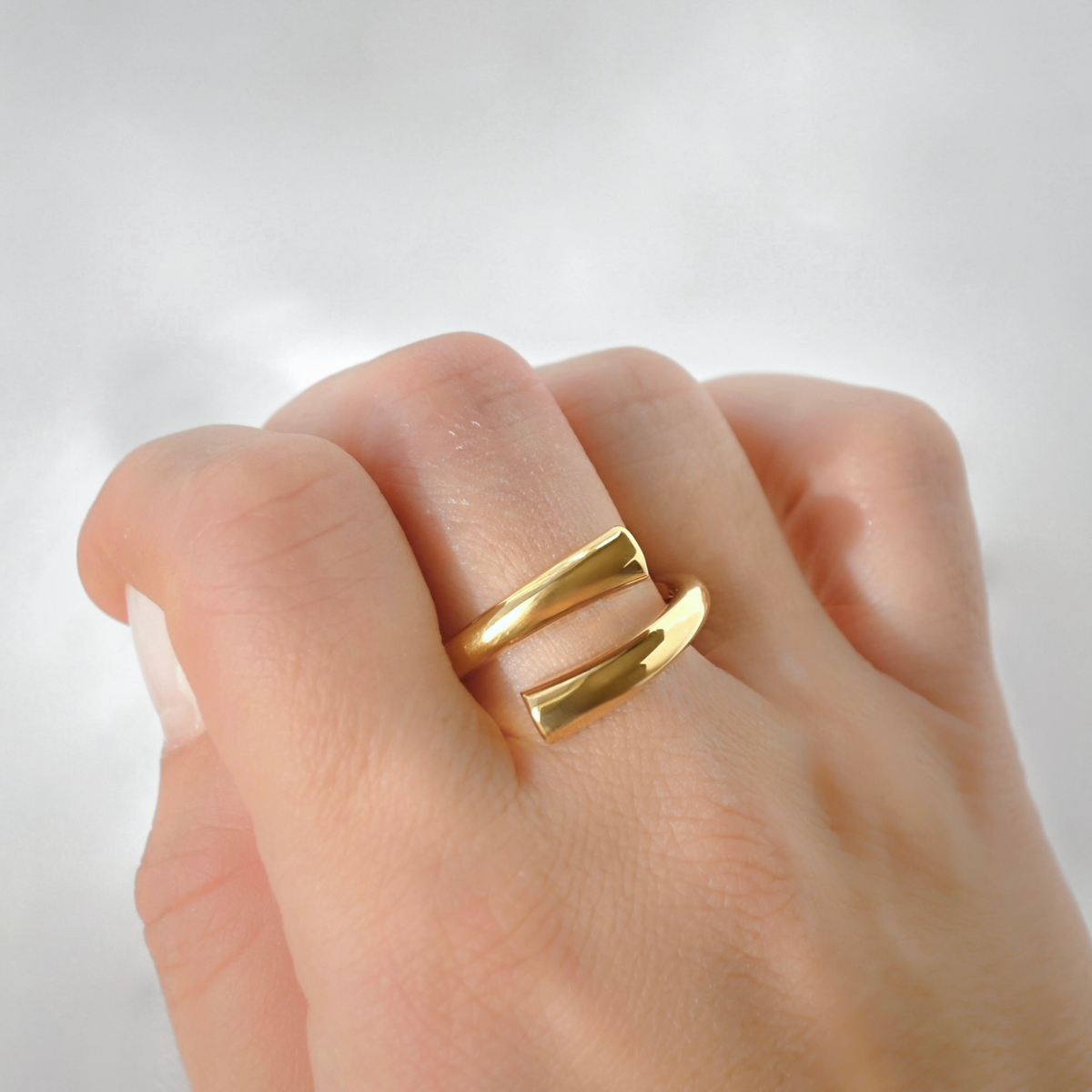 A close-up of a hand wearing a chunky gold ring with two parallel polished bands, photographed against a soft light gray background.