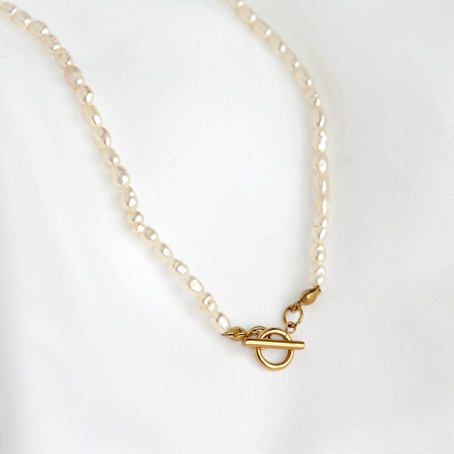A delicate necklace featuring small, irregular cream-colored elongated pearls, finished with a gold toggle clasp, displayed on a soft white fabric background.