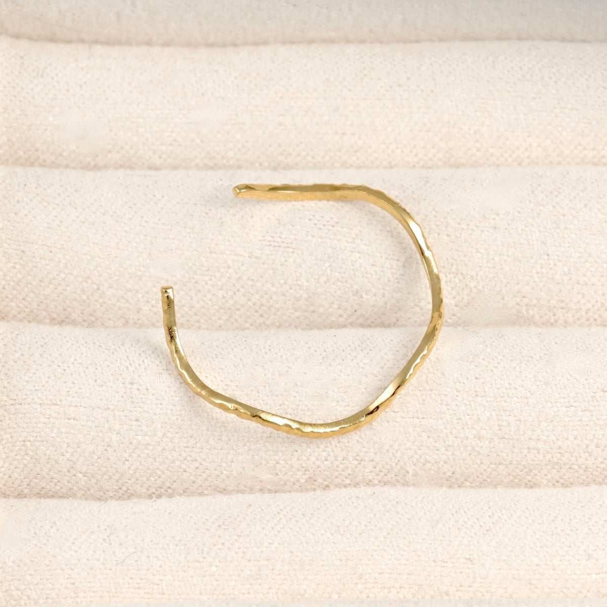 A single hammered gold open cuff ring displayed on a textured cream fabric surface.