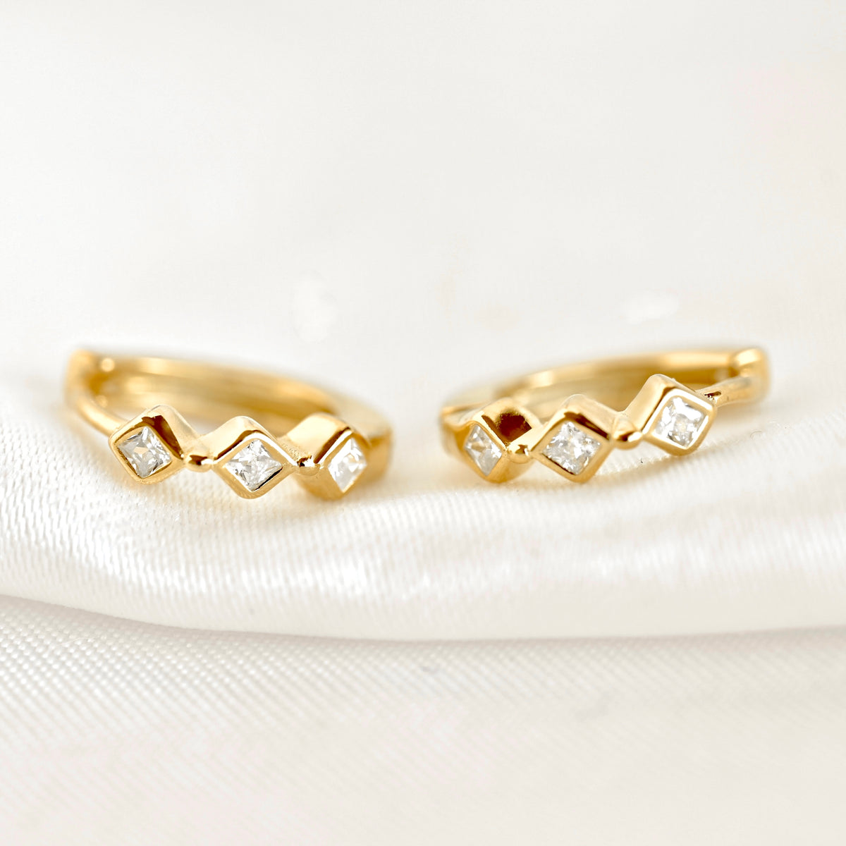 A pair of gold huggie hoop earrings with a zigzag design, each set with small clear square-cut gemstones, resting on a white textured fabric background.