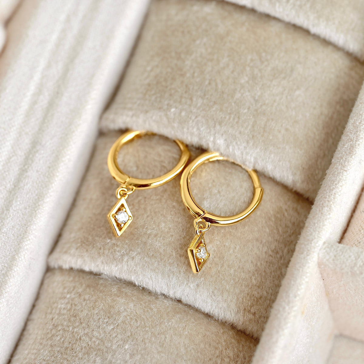 A pair of gold huggie hoop earrings with dangling geometric diamond-shaped charms set with small clear stones, displayed on a beige velvet jewelry box.