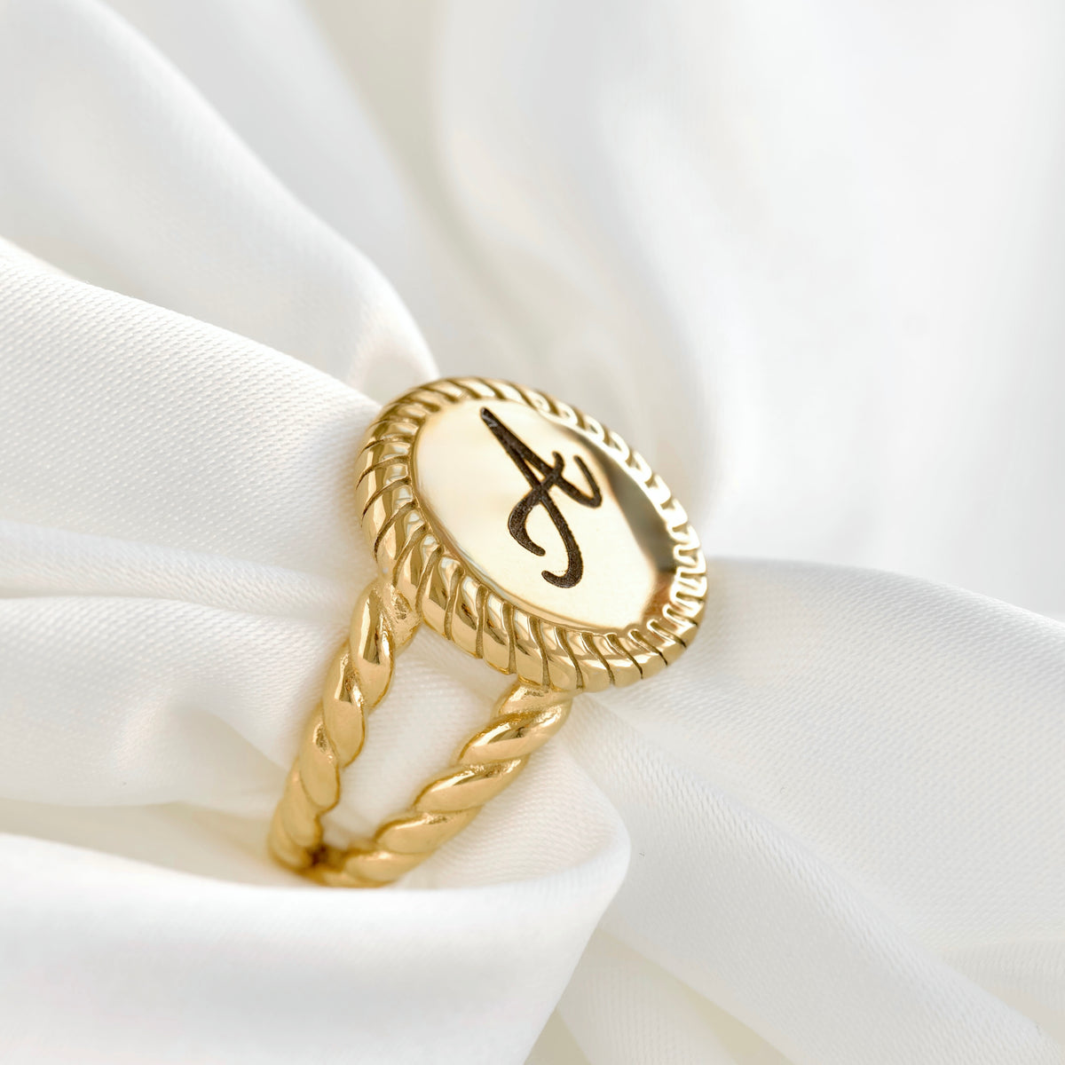 A gold ring with a twisted rope-style band and an oval face featuring a black monogram letter 'A' on a cream background, displayed on draped white fabric.