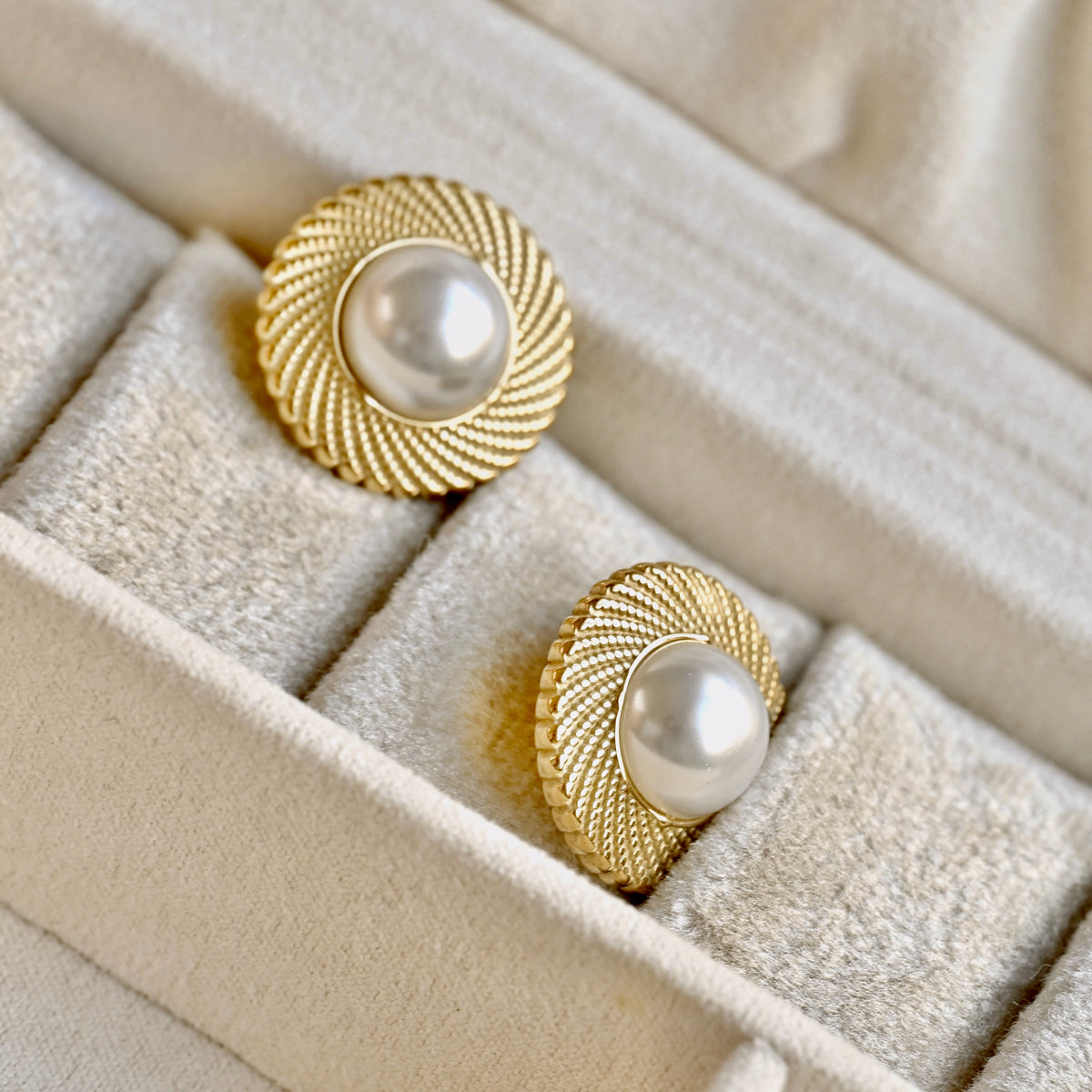 A pair of gold-toned round earrings with textured sunburst edges and white pearl centers, displayed in a beige velvet jewelry box.