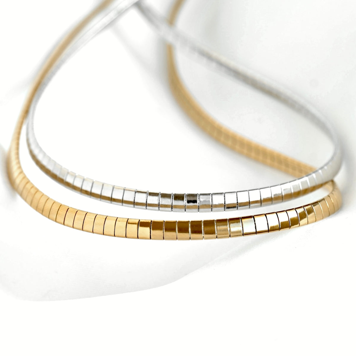 Two slim snake-bone style necklaces, one in polished yellow gold and one in polished silver, arranged in a layered loop on a clean white background.