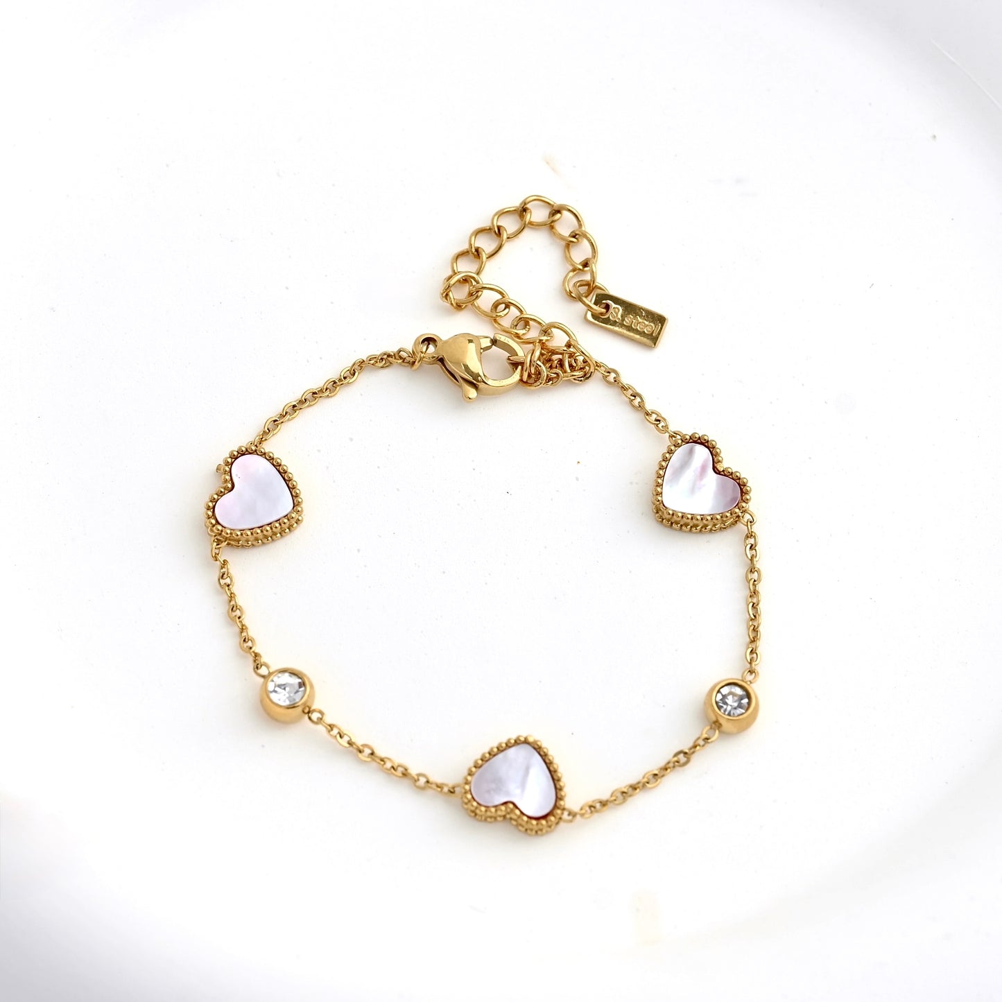A gold chain bracelet featuring three milgrain heart charms with white shell inlay,and two clear CZ accents, displayed on a clean white background.
