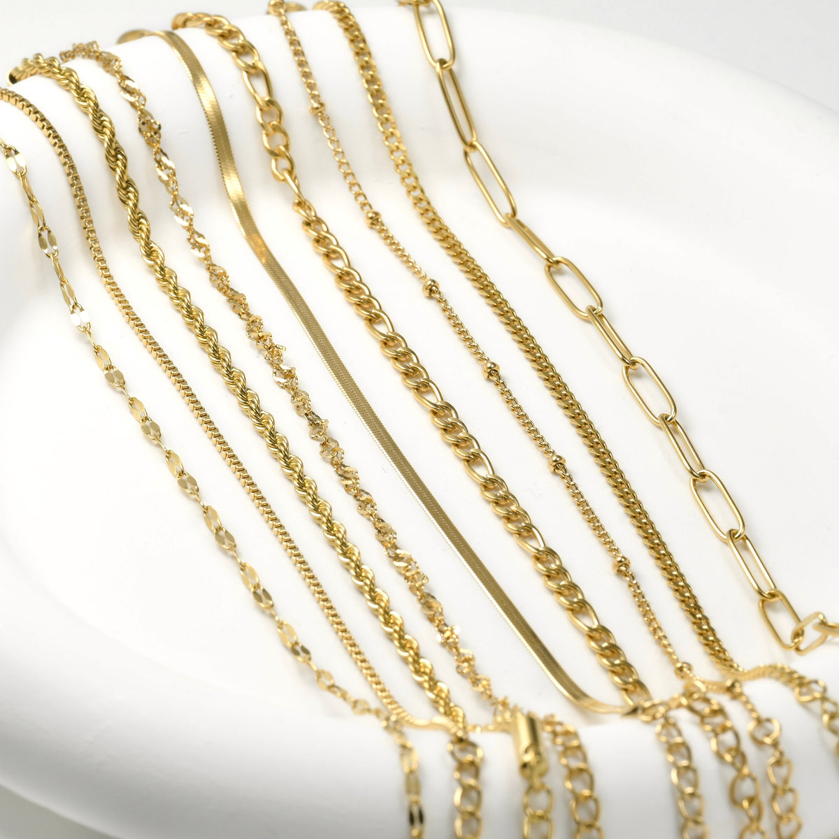 Collection of gold chain bracelets (mariner, box, rope, singapore, herringbone, figaro, satellite, 2mm curb and paperclip designs) on a white background.