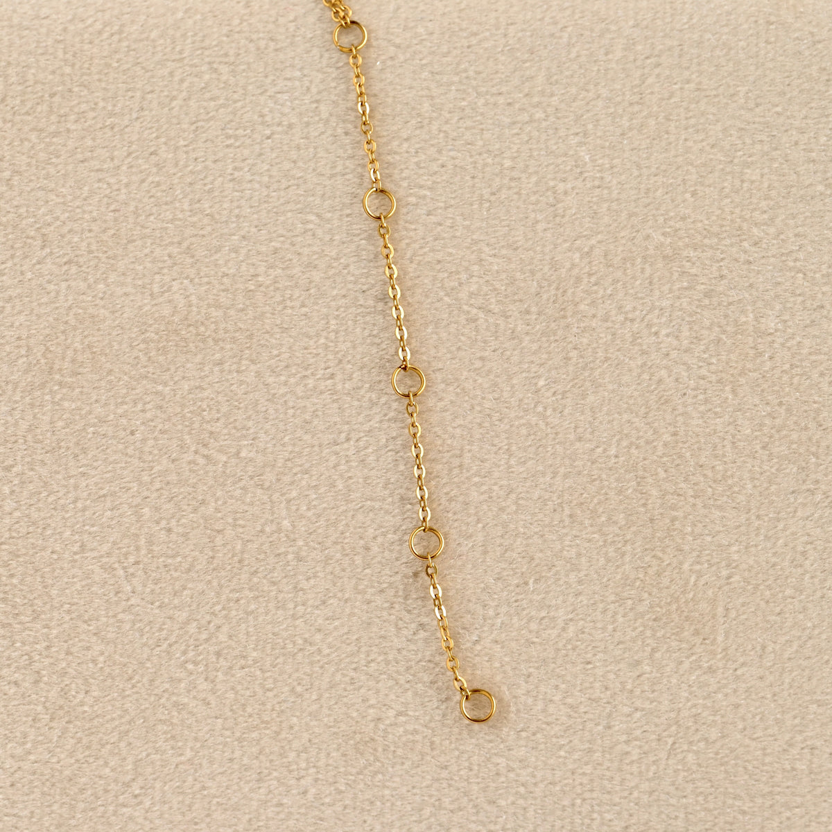 A close-up of a delicate gold chain extender with four small circular links and a drop loop, laid on a textured beige surface.