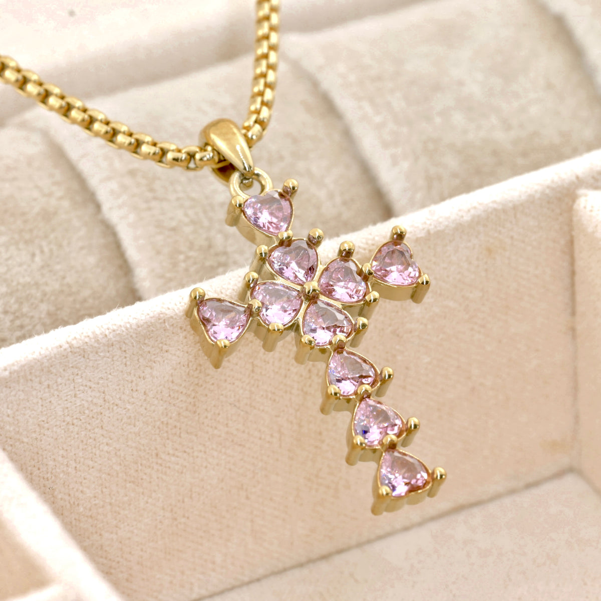 Gold cross necklace with pink heart shaped diamonds on a beige fabric background