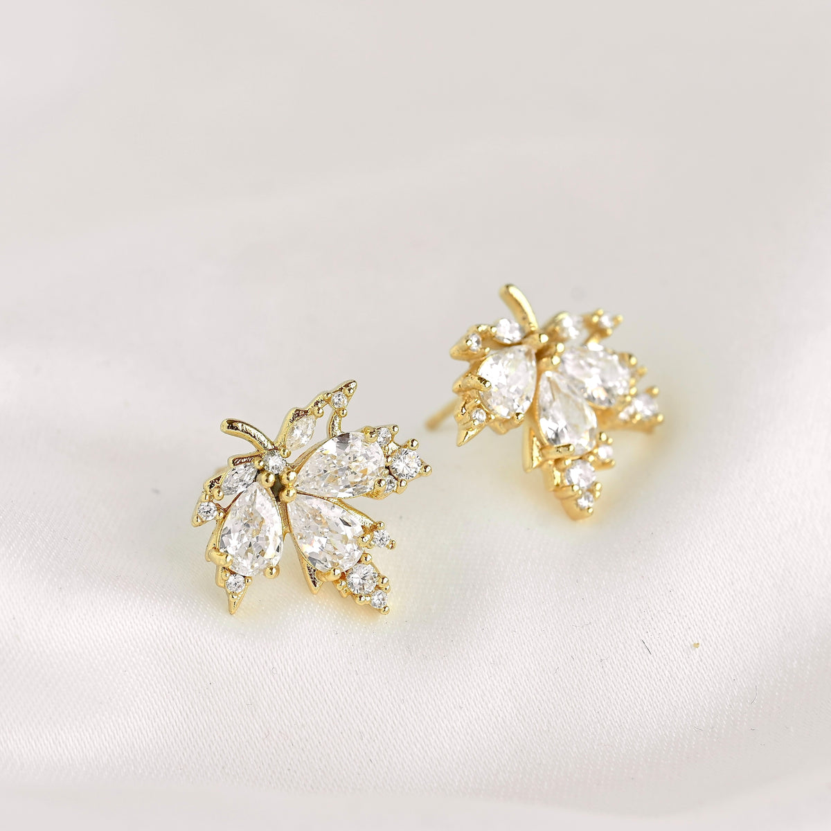 A pair of gold leaf-shaped stud earrings adorned with clear marquise and round crystals, displayed on a soft white fabric background.