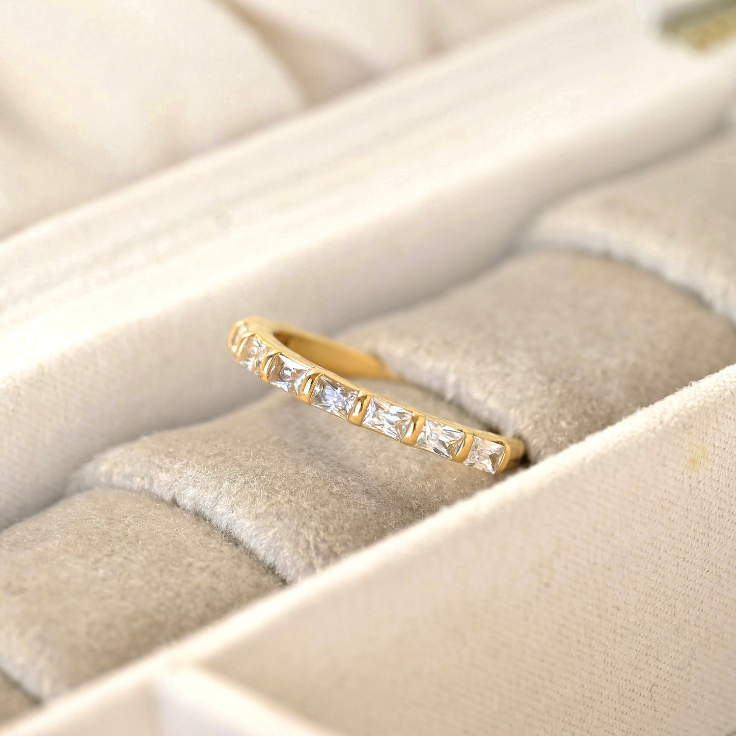 A yellow gold half eternity ring featuring a row of baguette-cut clear stones, displayed on a beige velvet ring box.