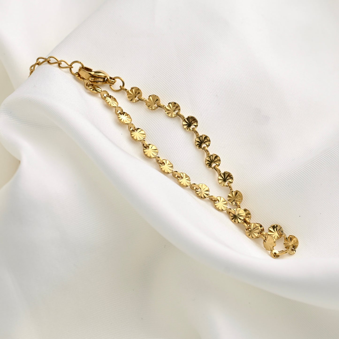 A gold chain bracelet with small round starburst links, displayed on draped white fabric.