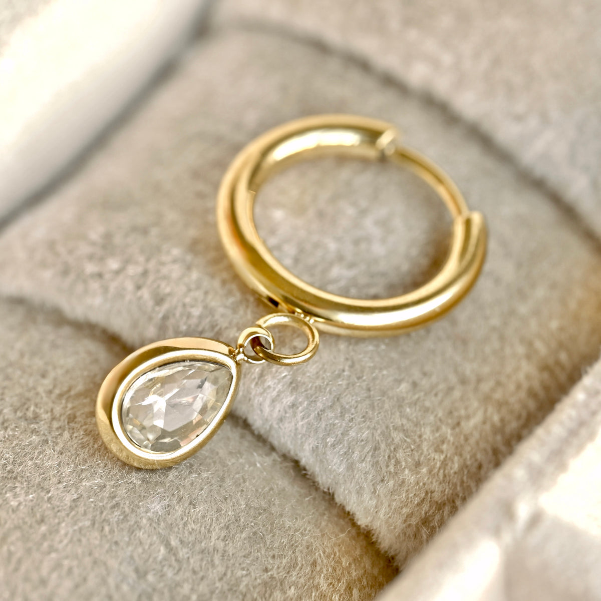A gold huggie hoop earring with a dangling teardrop-shaped clear faceted gemstone, displayed on a soft gray velvet jewelry box cushion.