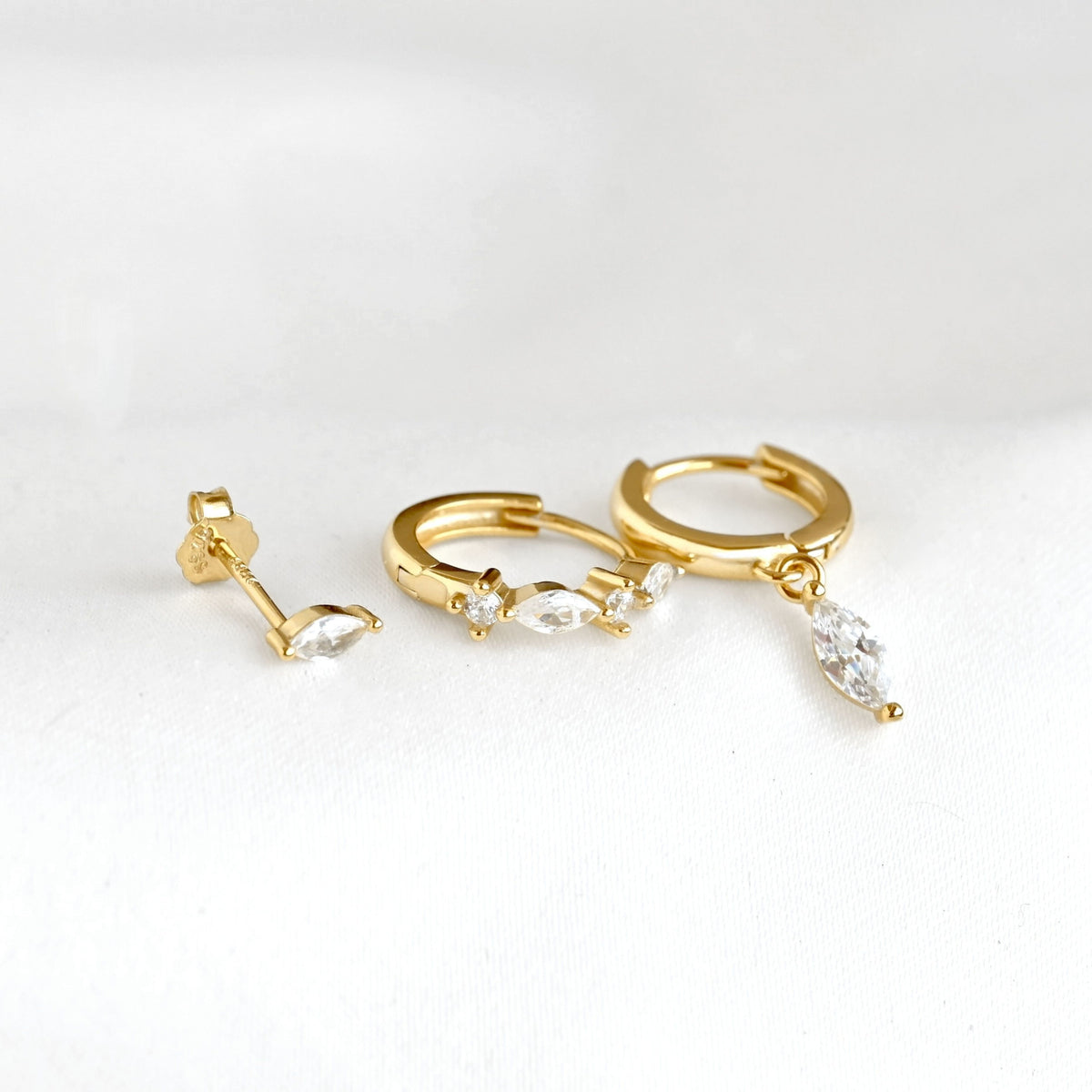 Gold huggie hoop earrings with marquise-cut clear crystals laid flat to display the dangling design, on a white background.