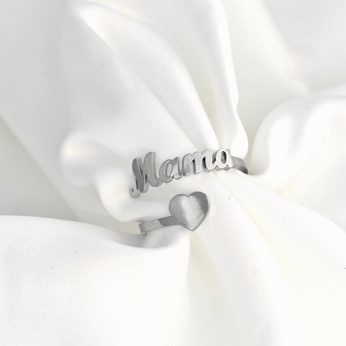 A silver ring with the word 'Mama' in cursive script and a small heart charm, displayed on soft white fabric.